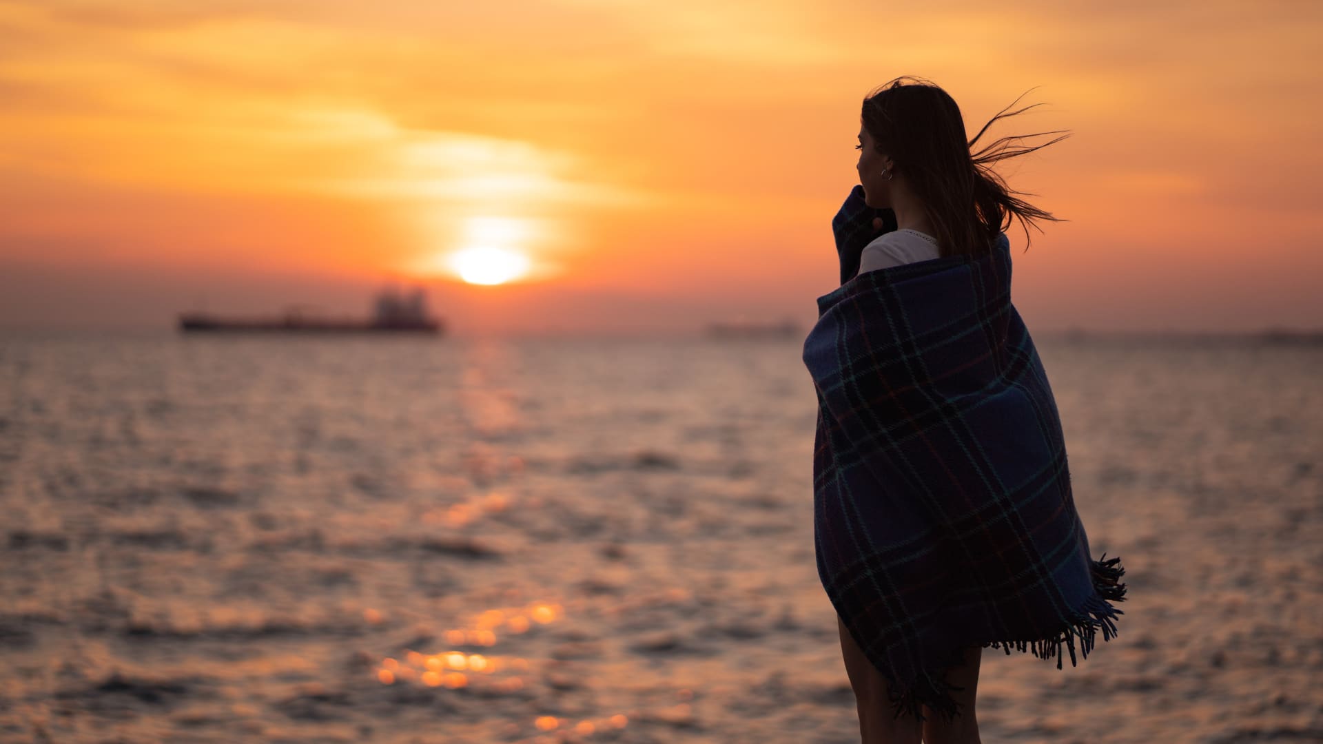 A woman wrapped in a blanket watches a sunset over the water