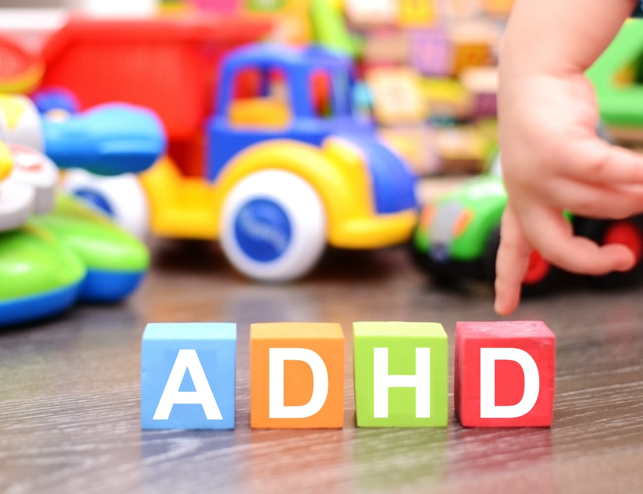 ADHD in the classroom.Image shows colorful toy blocks spelling out "ADHD" placed on a table with toy cars and trucks I'm the background. a child's hand is reaching towards the blocks, suggesting a playfully and child focused setting.